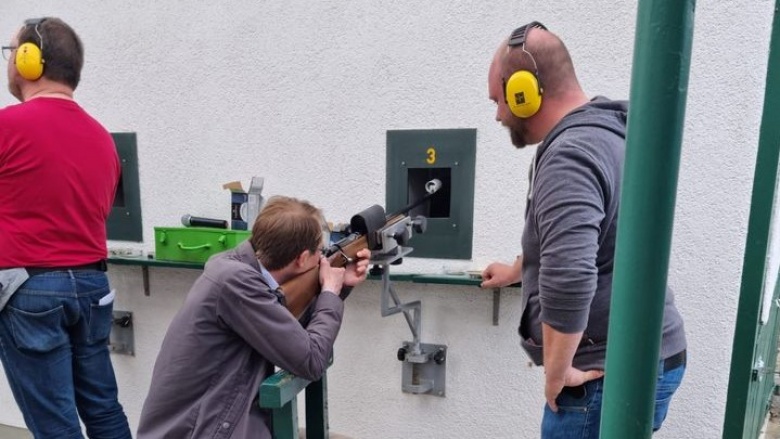 Dr. Christian Untrieser zielt mit einem Gewehr auf eine moderne Zielvorrichtung. Rechts und links von ihm stehen zwei Männer mit Gehörschutz und beaufsichtigen das Schießen.