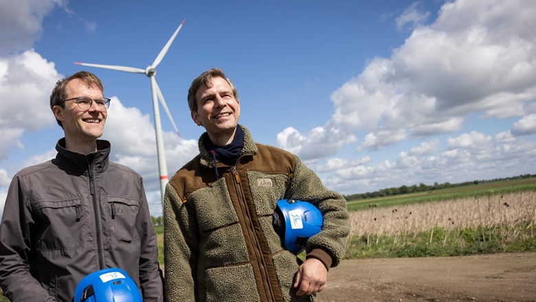 Hoch hinaus für erneuerbare Energien und Bürgerbeteiligung Dr. Christian Untrieser (links) und Stephan Wolter (rechts) stehen vor einem Windrad. Sie haben blau Schutzhelme unter dem Arm und lächeln.