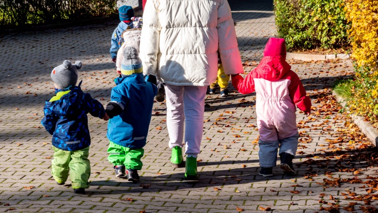 Erzieherin geht mit Kindern draußen spazieren. Die vier Kinder tragen Schneeanzüge, Gummistiefel und Mützen oder Kapuzen. Die Sonne scheint auf ihren Rücken.