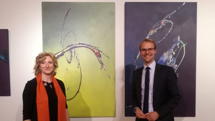Künstlerin Viola von Dyk und Dr. Christian Untrieser stehen lächelnd vor zwei großformatigen Malereien. Diese zeigen Wasserfontänen auf grünem und dunkelblauem Grund.