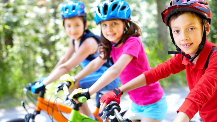 drei Kinder in bunter Kleidung und mit Schutzhelm auf Fahrrädern